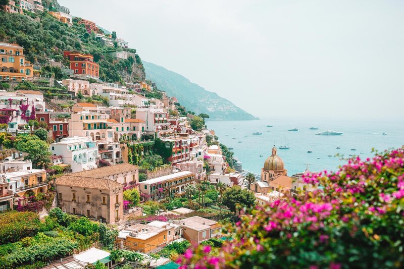 Scenic view of a coastal town with colorful buildings on a hillside overlooking the sea. Boats are scattered across the water. In the foreground, vibrant pink flowers add contrast to the lush greenery and architecture, creating a perfect backdrop for luxurious Caribbean getaways.