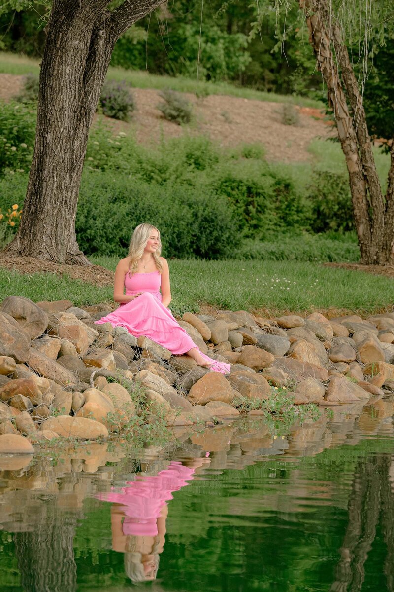 graduates refelction shows in the water as she sits on the rocks wearing a long pink dress