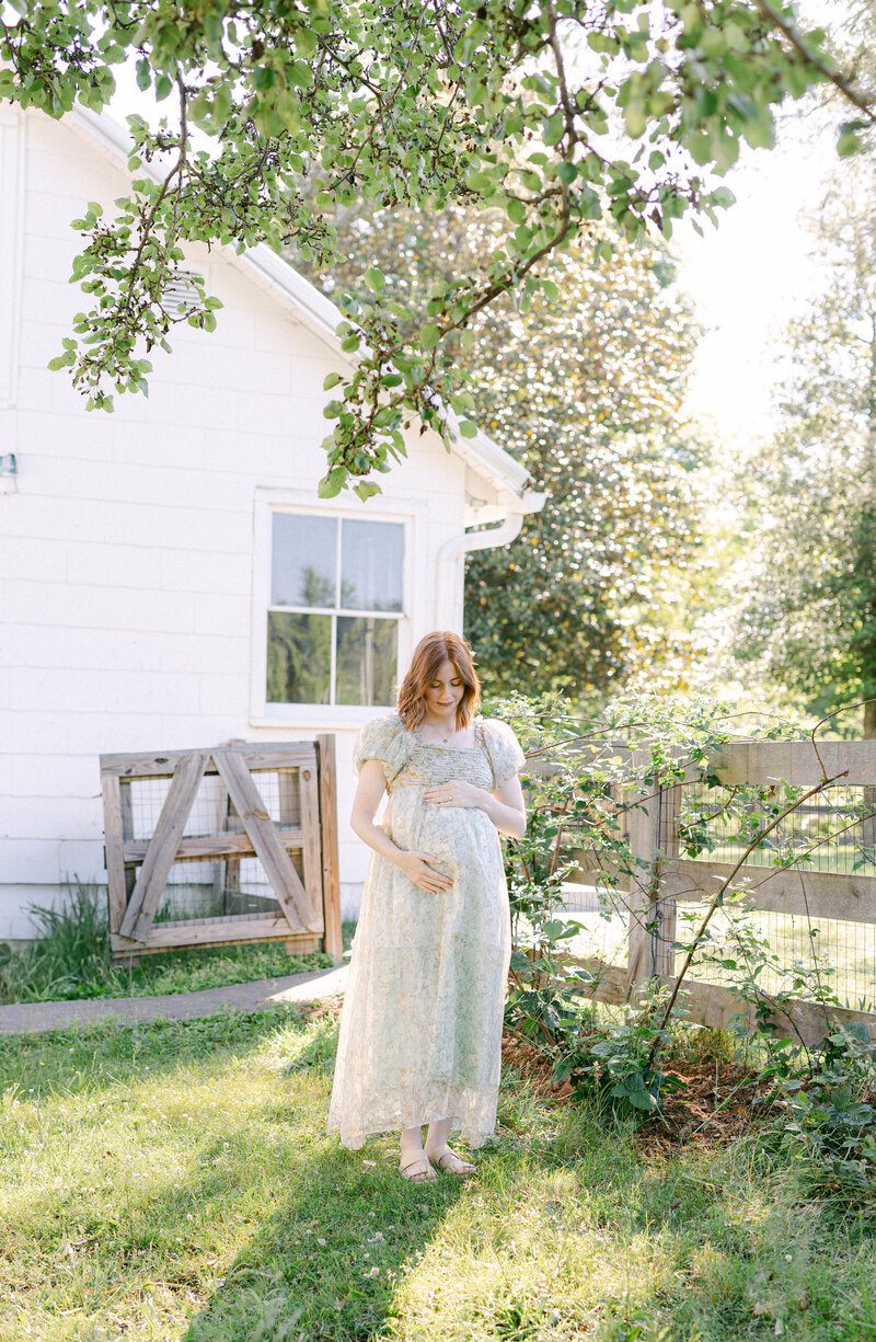 knoxville tennessee newborn photographer mom holding belly bump while wearing a sheer green dress being sunlit from behind surrounded by nature