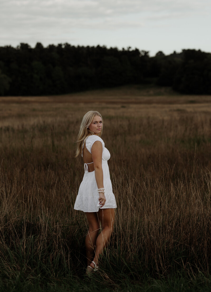 A woman in a white dress stands in a grassy field, gazing over her shoulder. The scene captures the essence of portrait photography New England, with a dense tree line under a cloudy sky setting an atmospheric backdrop.
