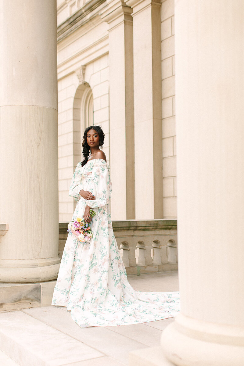 A beautiful black bride stands alone with her bouquet near the classical columns of Lansing's Capitol Building. She has glowing skin and a soft beautiful look.