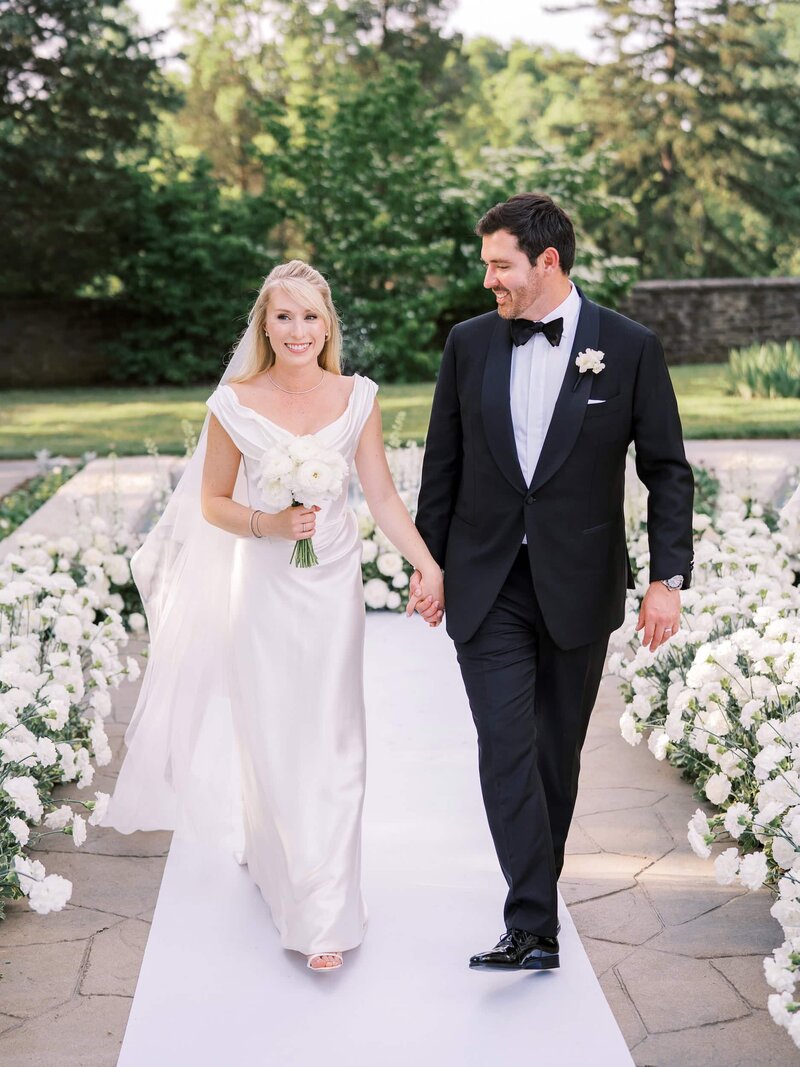 Bride and groom walking hand in hand down a white aisle lined with white flowers.