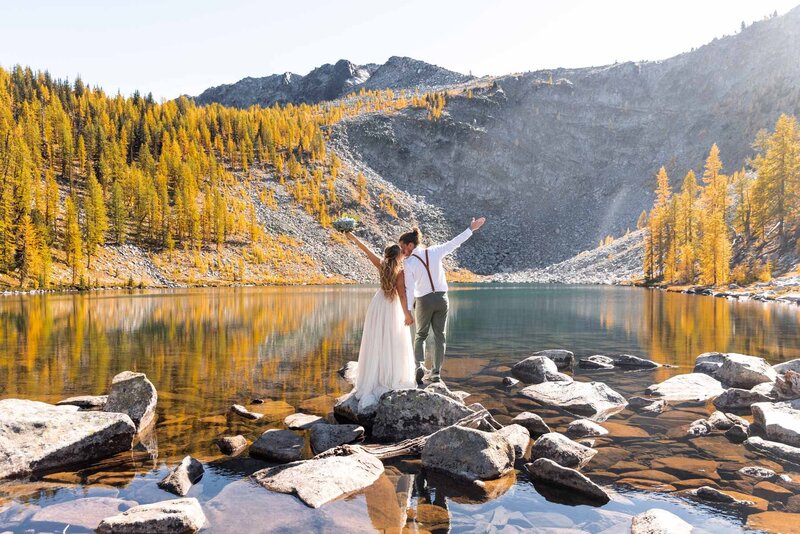 A couple celebrating with arms up and kissing while standing on rocks in the middle of the lake with lots of golden larches surrounding them