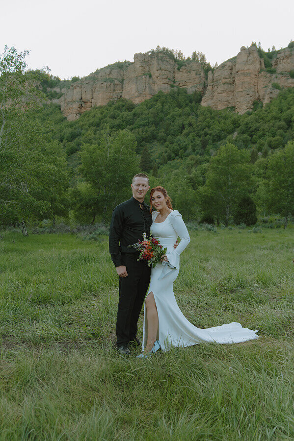 Bridal portraits of a bride and groom. They are standing next to one another in a grass field with cliffs behind them.