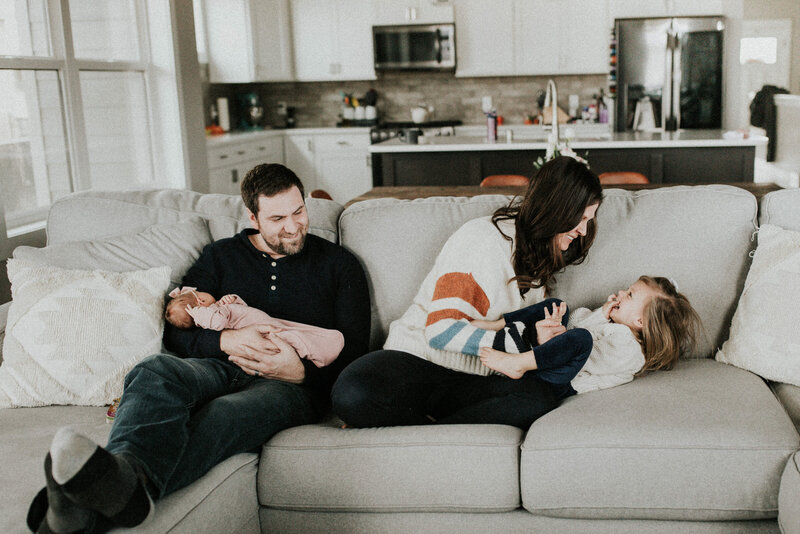 A man holds a baby as a woman tickles their daughter all while sitting on the couch