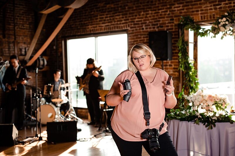 Funny behind the scenes photo of photographer working a real wedding, she's holding camera gear and making a funny wink face at the camera