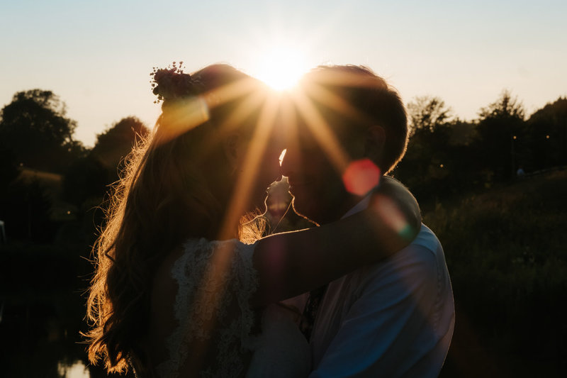 Bride and groom embrace in the sunset at Hadsham Farm weddings. Orange sun shines over their heads making beautiful star shaped beams