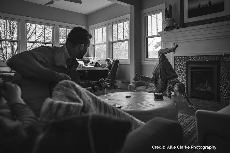 A black-and-white image of a family inside a cozy living room. A man plays the guitar while a child performs a handstand near the fireplace, and another child sits in the background, reading.