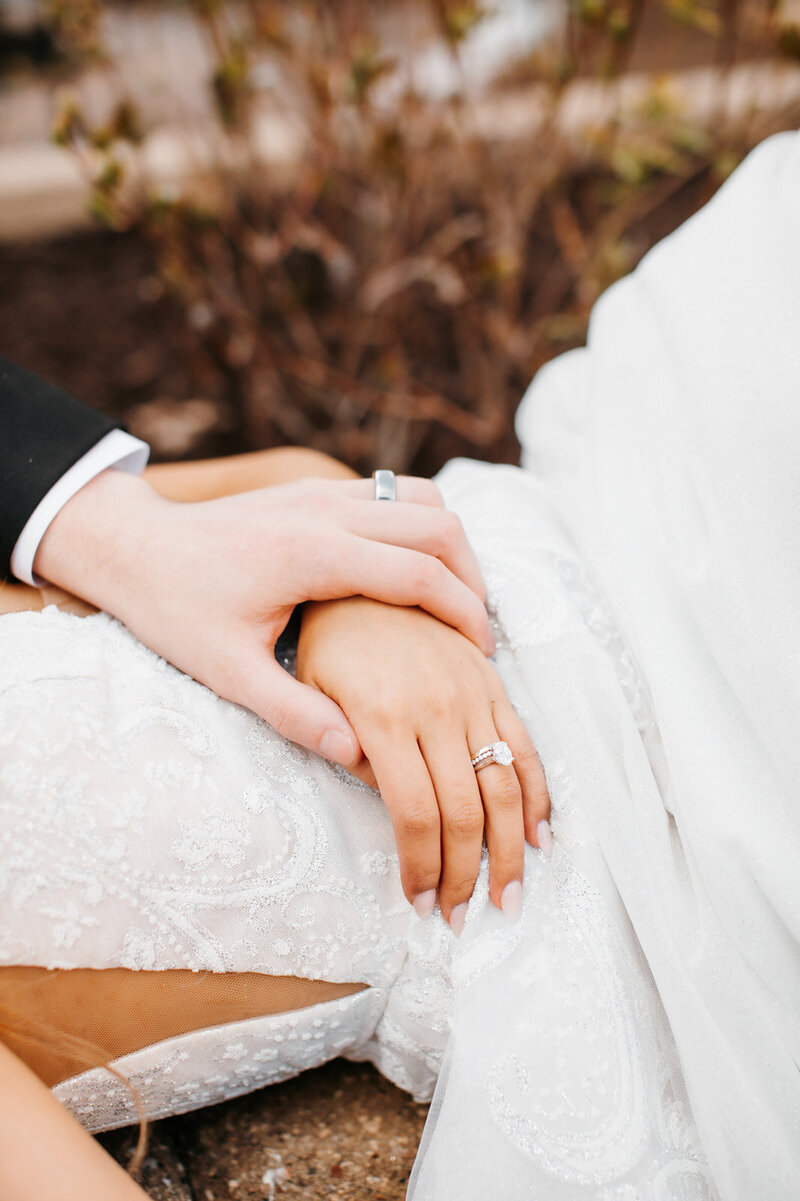 Close up photo of bride and grooms' hands together resting on top of the bride's dress