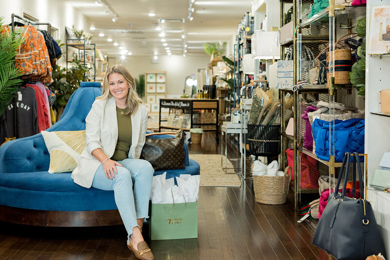 Smiling woman sits on a plush blue chaise inside a stylish boutique in Columbia, SC, surrounded by curated fashion, accessories, and home décor. A shopping bag from Icon on Taylor rests beside her, highlighting a luxury retail experience. Small business branding photography in Columbia, SC, capturing upscale shopping, personal branding, and local business marketing.