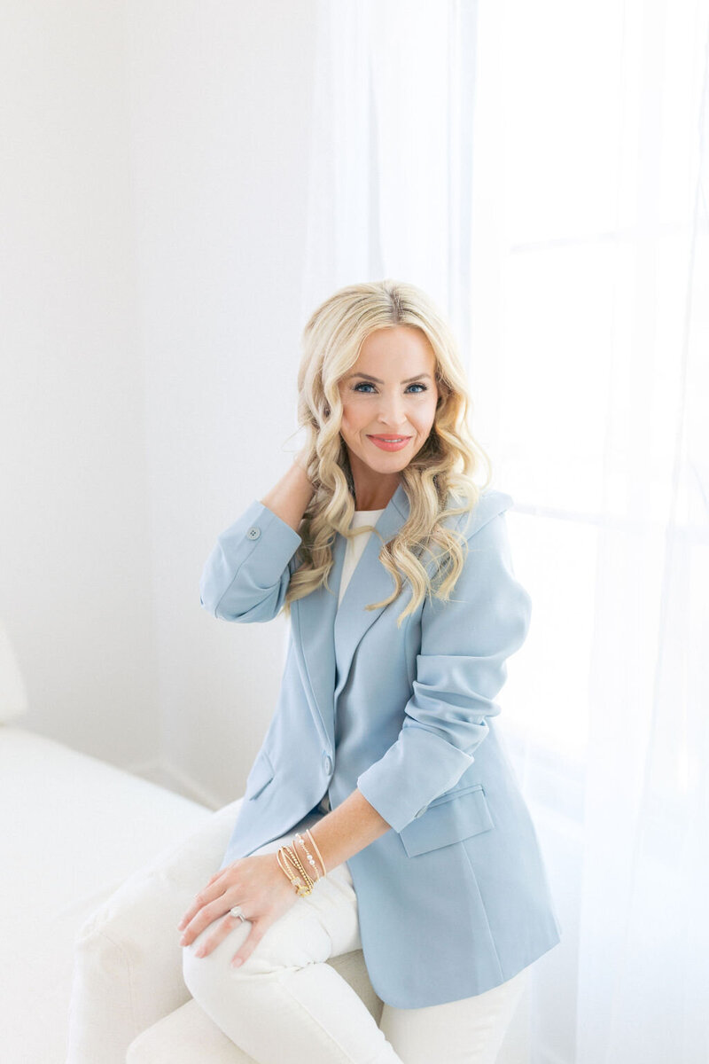 Woman with blonde hair and blue jacket sits on white couch and holds hair with right hand