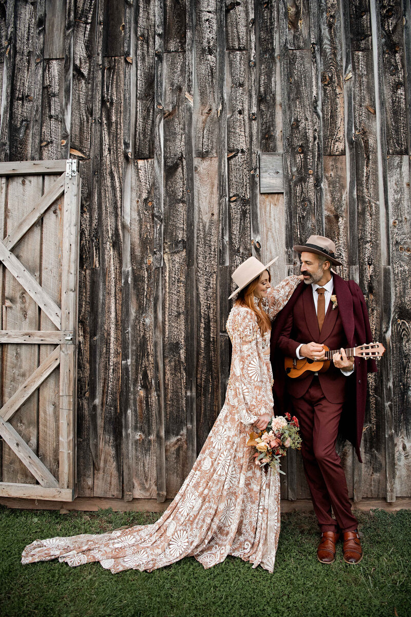 A groom in a burgundy suit plays and sings to a bride in a hat and detailed printed gown.
