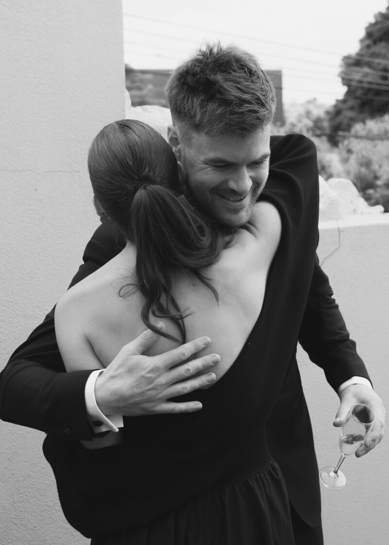 A black-and-white close-up photograph capturing an intimate moment between the groom and a guest at his wedding. The groom, dressed in a sharp black tuxedo, is embracing the guest warmly, smiling broadly as he holds a half-full champagne glass in one hand. His hand is gently resting on the guest’s back, who is wearing a dark dress with an open back and has her hair tied back in a low ponytail. The backdrop is simple, featuring an outdoor setting with a light-colored wall and soft, blurred trees in the background. The candid nature of the image captures the joy and emotion of the day, focusing on the connection between the two individuals, exuding warmth and happiness. The timeless black-and-white filter adds a classic, elegant touch to the scene, highlighting the textures and expressions.