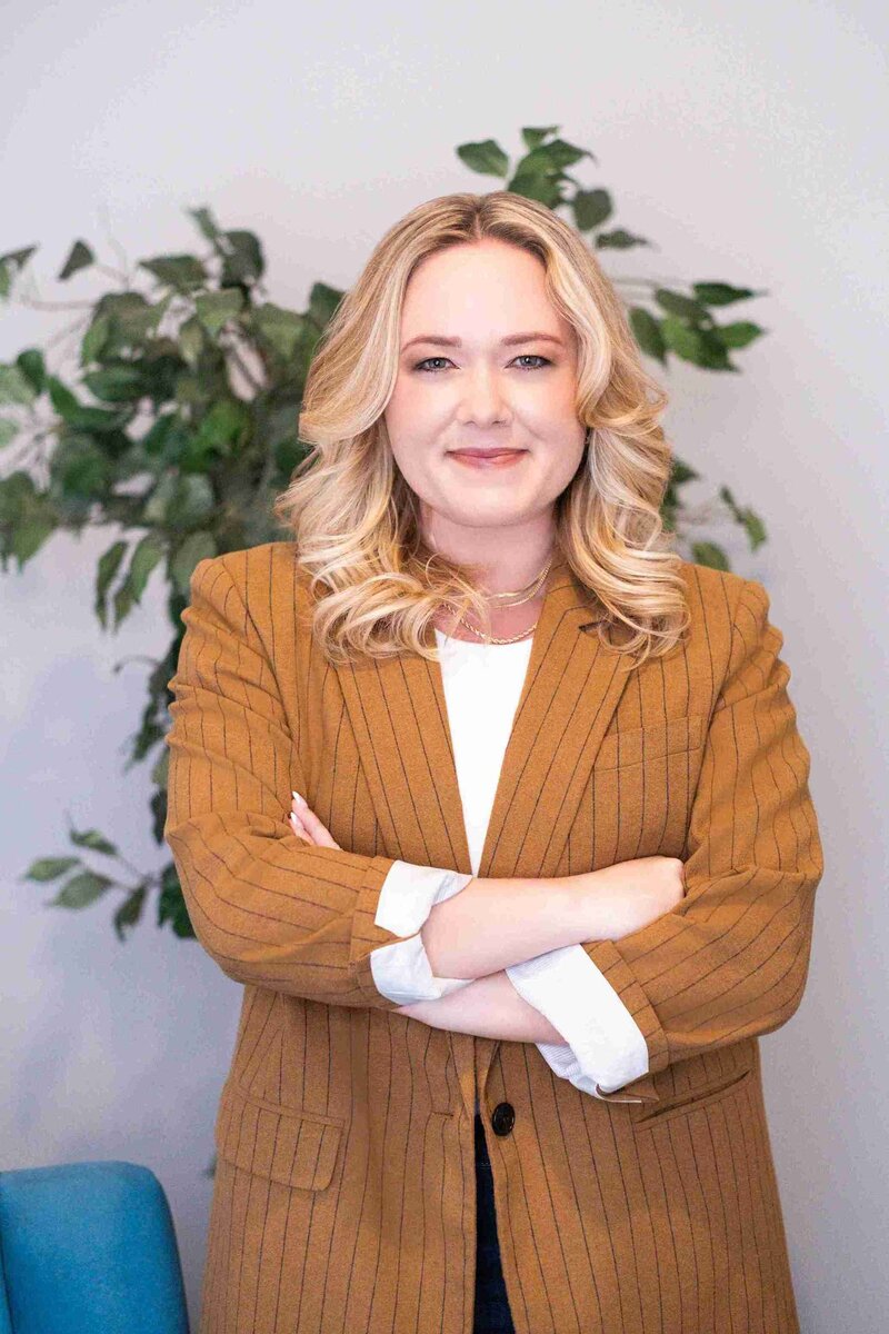 A woman in a suit and white shirt stands confidently by a blue chair, representing her role as a New York therapist media consultant.