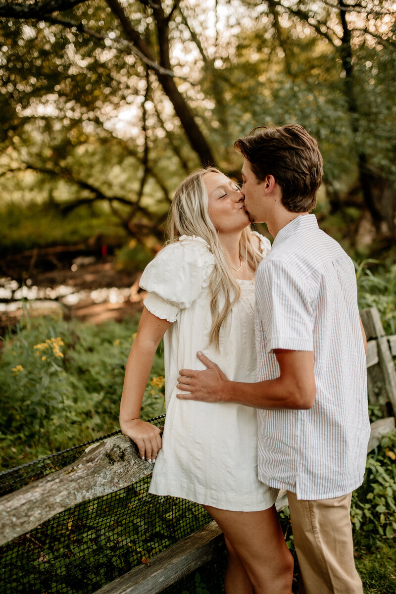 Engaged couple shares a kiss along an old fence in a serene forest as a babbling brook flows nearby.
