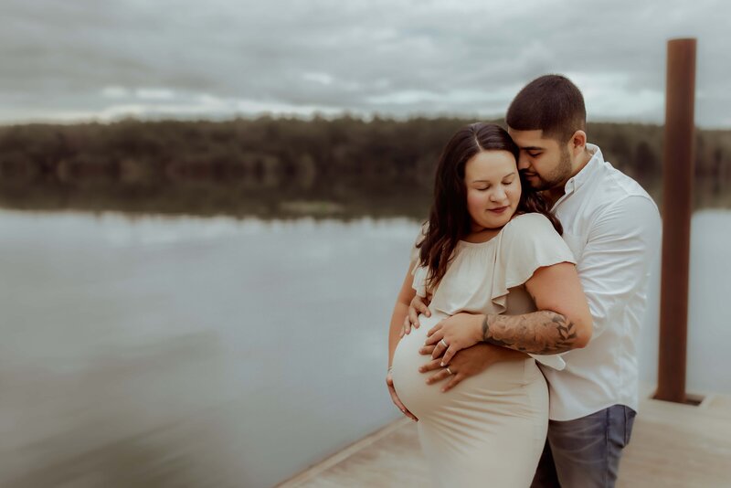 A pregnant couple embracing on a dock in front of a lake.