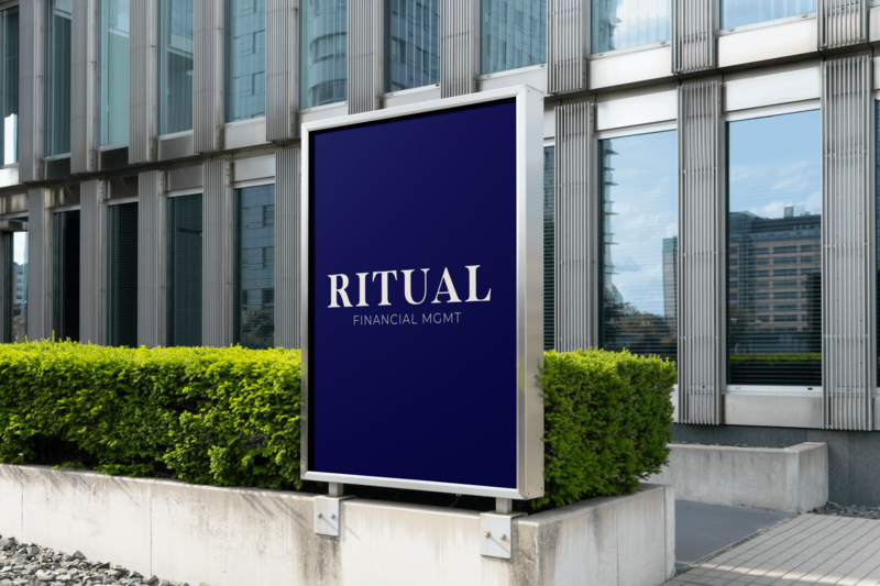 A modern office building with a blue sign reading "RITUAL FINANCIAL MGMT" in front of green shrubs.