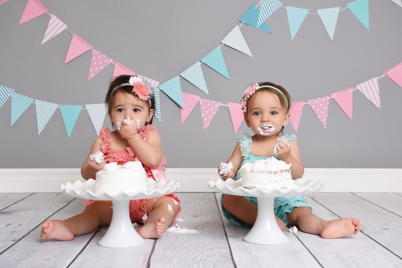 Twin baby girls enjoying their cake smash session, one in a coral dress with a pink flower headband and the other in a turquoise dress with a matching flower headband, with pastel bunting decorations in the background.