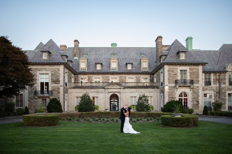 New England Wedding Photographer Pricing | Kelsey Sheehan Photography Timeless Rhode Island Weddings | A couple in wedding attire embraces on a manicured lawn before a grand, historic mansion. The scene conveys romance and elegance under a clear sky.