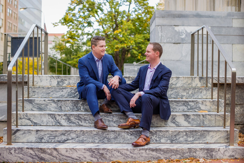 Two grooms holding hands on the historic steps of the Second Bank in Philadelphia, celebrating their wedding day.