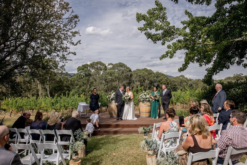 A micro wedding cermony with the bride and groom stnading on a raised platform, and the guests sitting in white chairs on the grass.