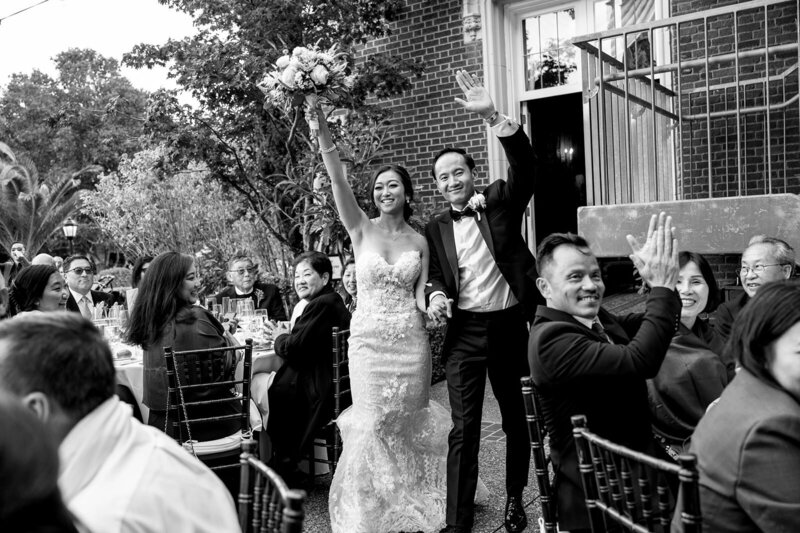 Professional San Francisco Bay Area wedding photography documenting authentic celebration moment as bride in beaded mermaid dress and groom in black tuxedo raise bouquet and arms in victory at Kohl Mansion reception with cheering guests and historic brick architecture, captured in timeless black and white