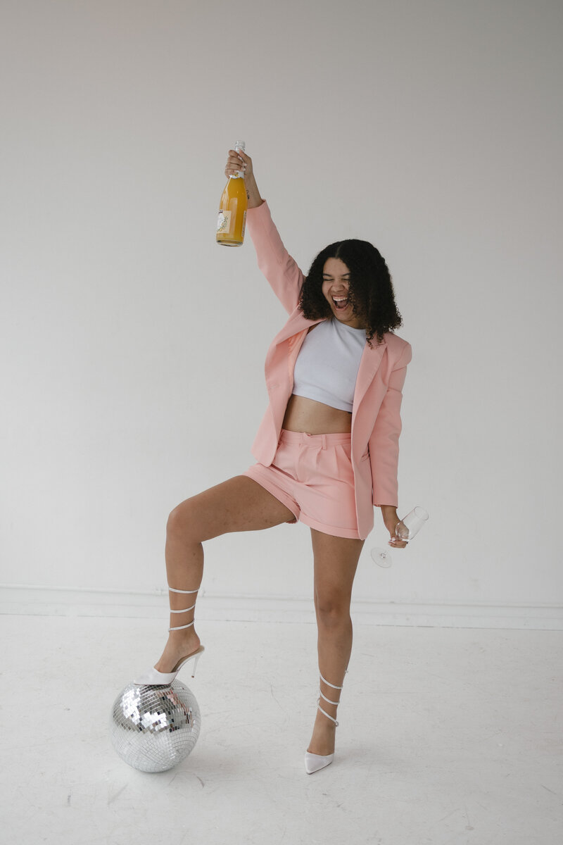 Photo of Catryce holding a bottle of mimosa in a pink jumpsuit with her white heels on a disco ball