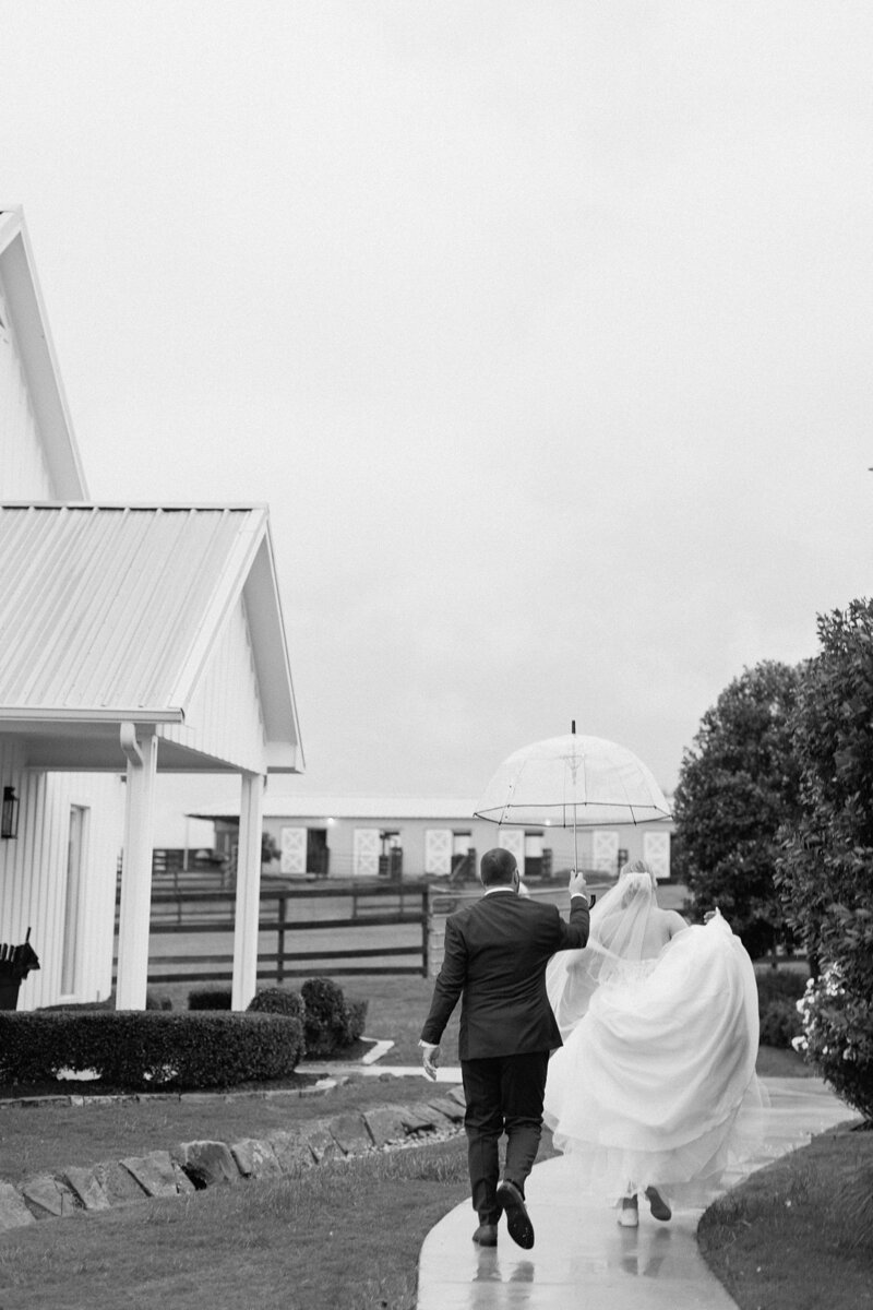 Black and white of bride and groom making the most out of their wedding day on a rainy day at The Farmhouse. The bride is holding up her dress and walking towards the chapel while the groom holds a clear umbrella over her head.