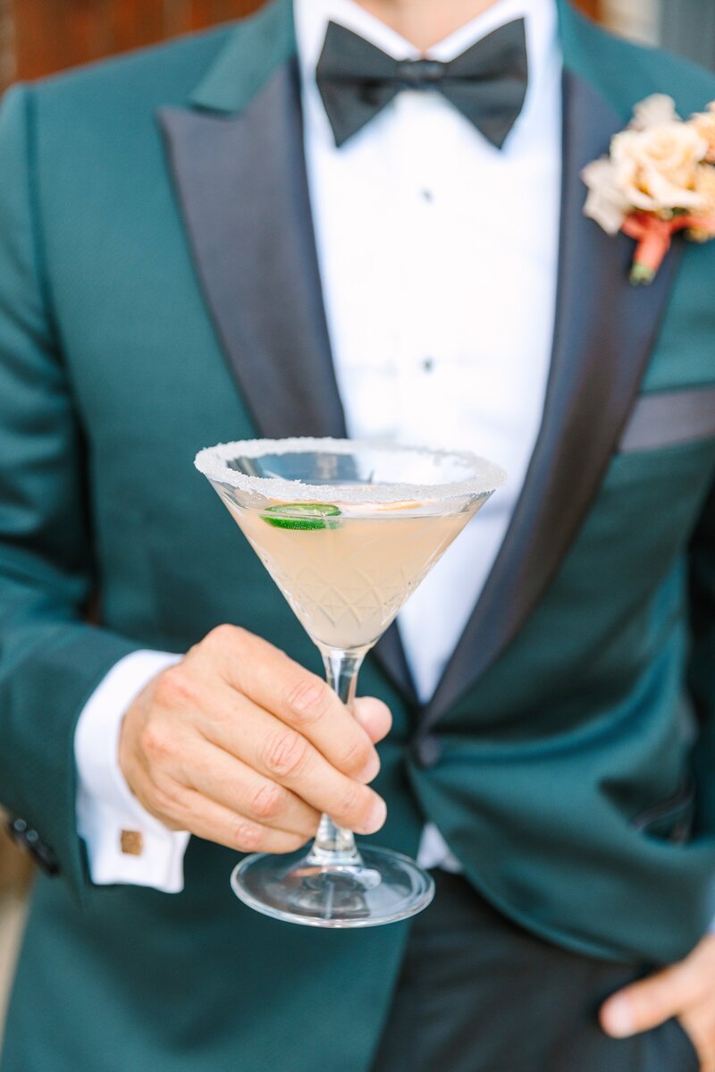 groom wearing dark green tuxedo jacket holds a martini