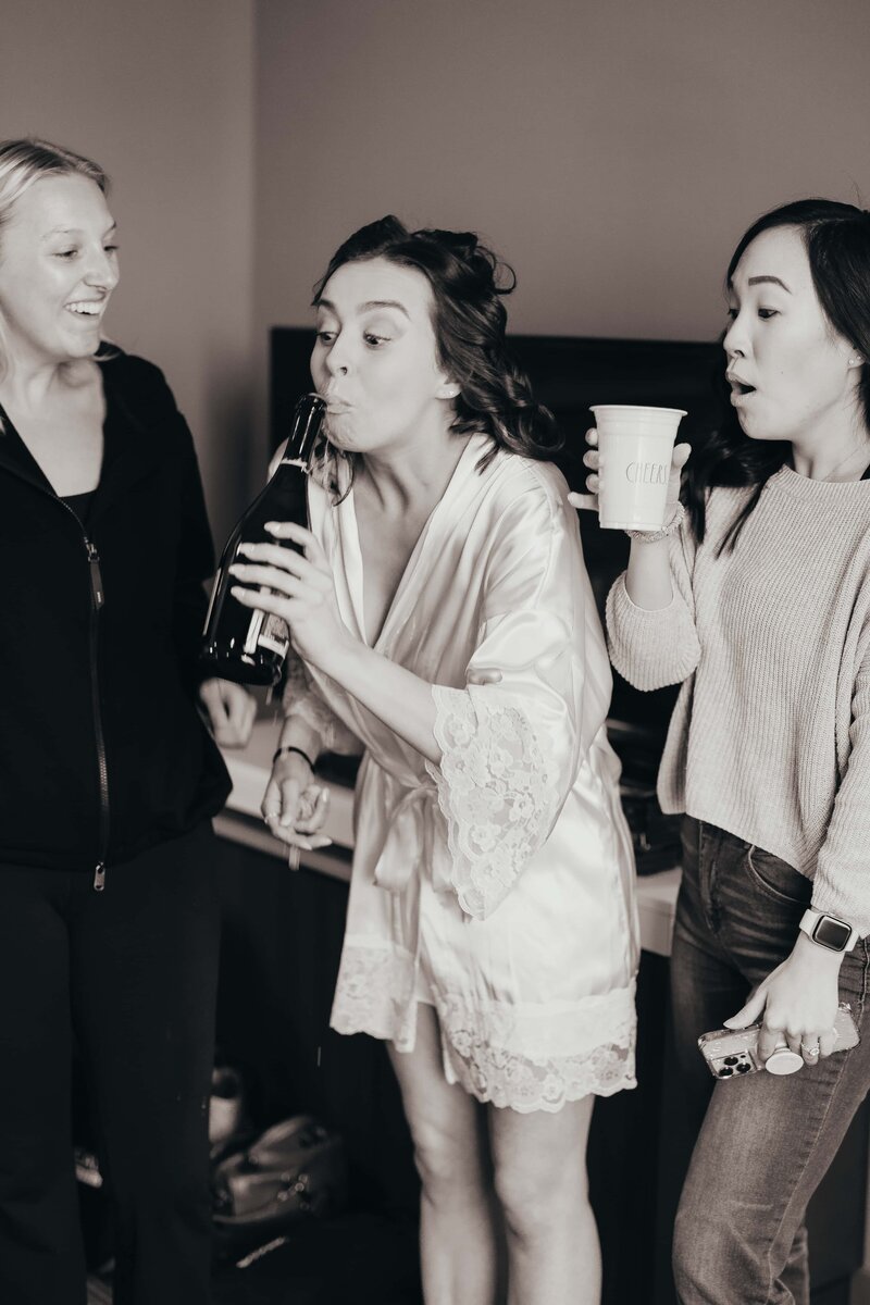 A woman in a white robe sips from a bottle while laughing with two friends in a casual indoor setting during an Iowa wedding planning session.