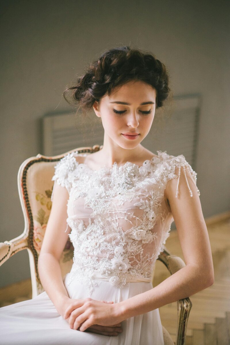 Bride wearing an elegant lace wedding gown with intricate floral embroidery and beaded details, sitting gracefully on a vintage chair