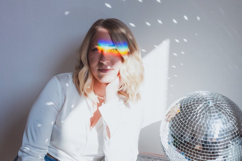 Woman with short, blonde, wavy hair softly smiles resting her head on her hand with reflected rainbow and discoball shine on her face