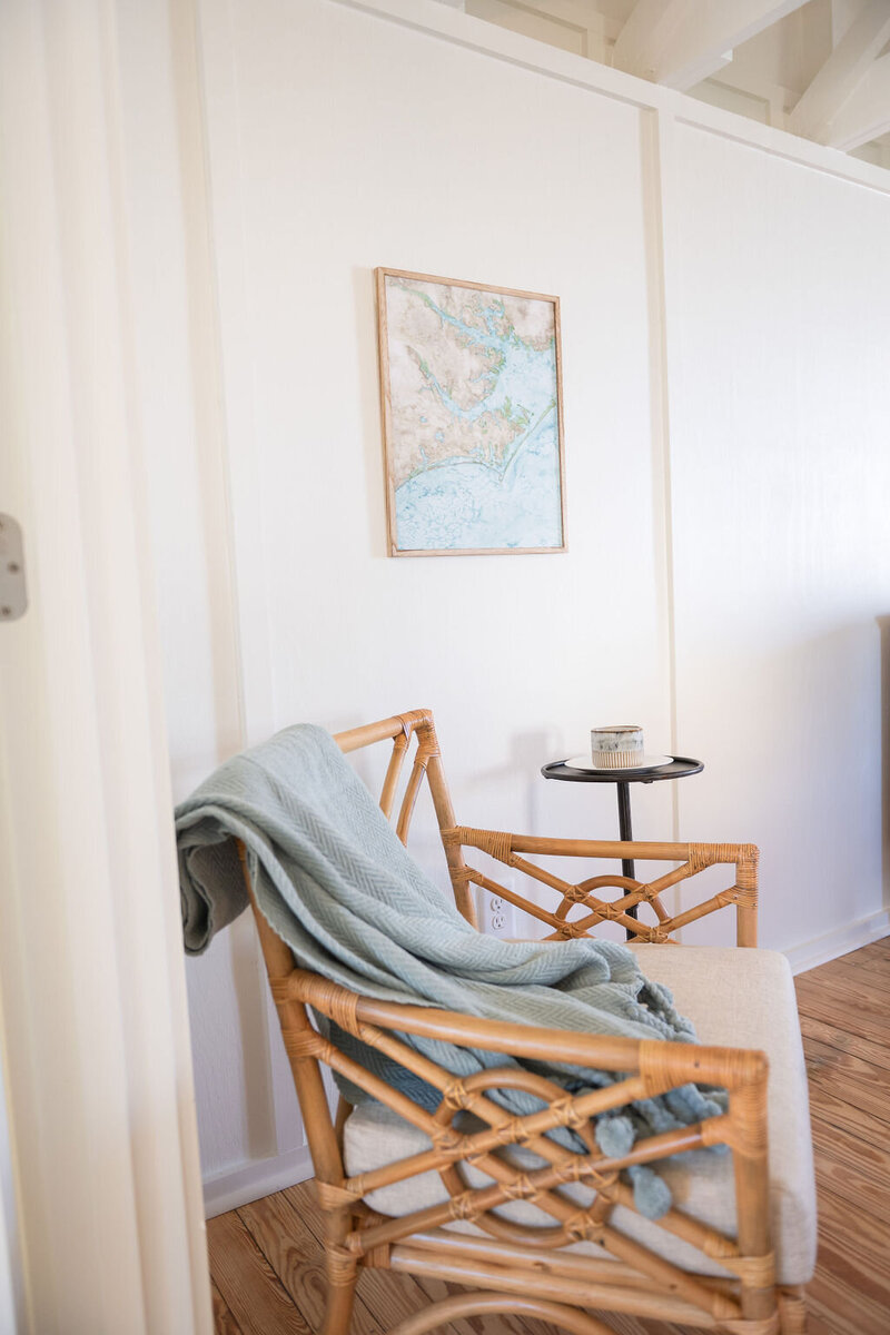 a boho rattan chair with a framed canvas of a watercolor painting featuring a coastal map with a candle on the round coffee table