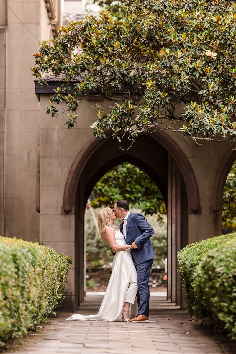 Bride and groom sharing a romantic kiss under a historic archway surrounded by greenery in Savannah, GA, during their elopement.