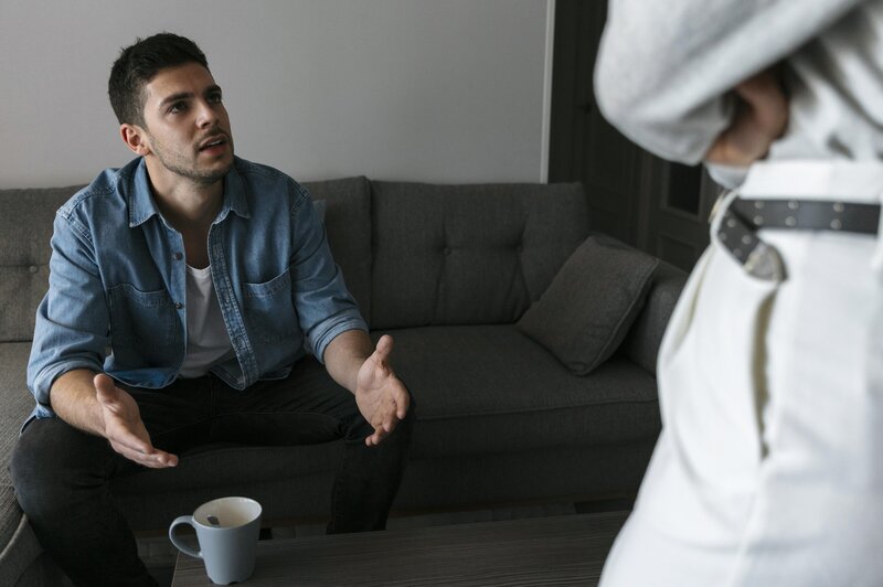 A man in a denim shirt sits on a gray couch with his hands open in a frustrated gesture, speaking to someone standing in front of him whose torso is partially visible.