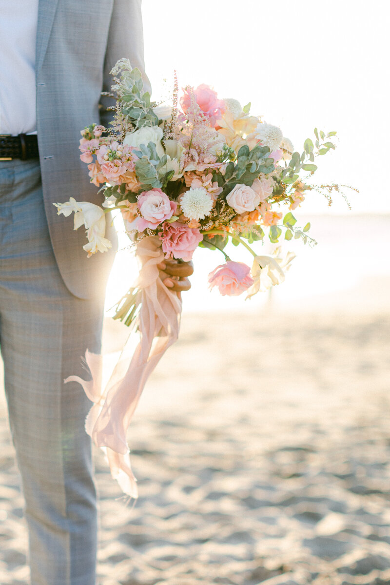 Groom holds wedding bouquet on the beach as ribbons blow in the breeze