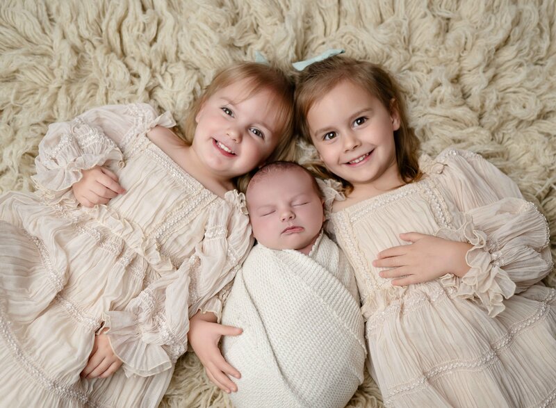Big sisters laying on fluffy rug with newborn baby brother between them. Photographed by Atlanta newborn photographer.
