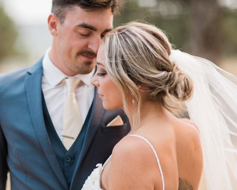 Image features a bride with a low messy up do and tulle veil flowing in the wind looking over her shoulder, while her groom in his blue suit and cream tie nuzzles his nose up to her head.
