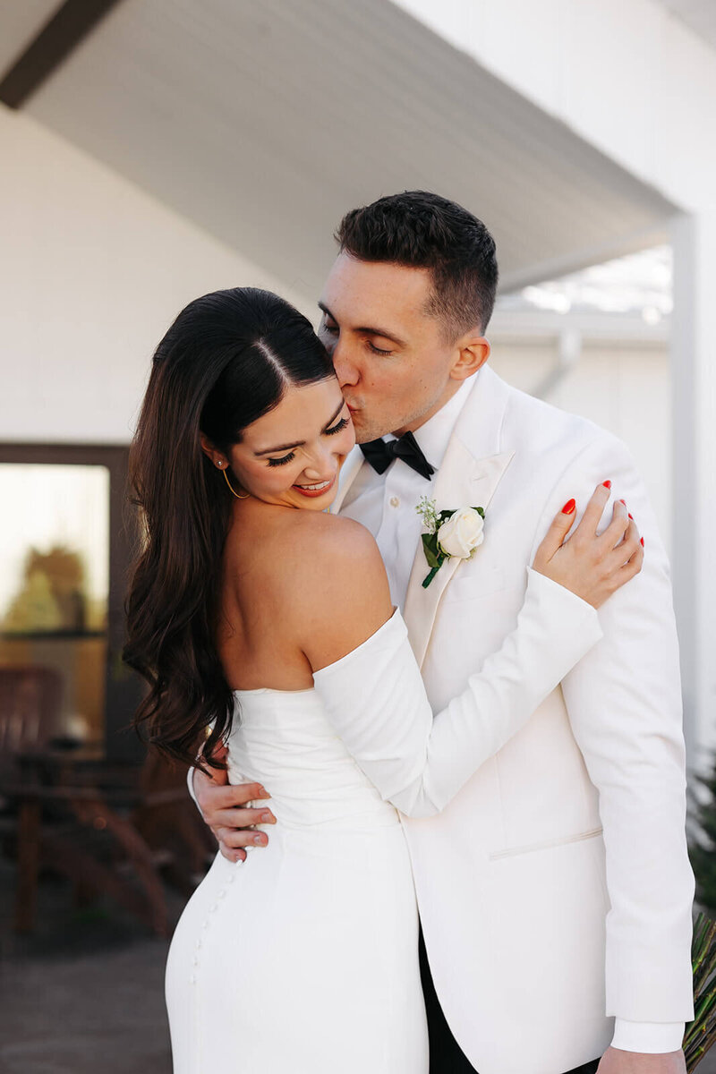 Groom in white tuxedo kisses bride's cheek at luxury Minnesota wedding