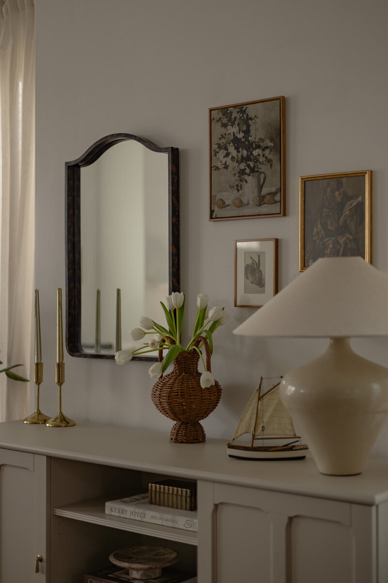 A detail image of a styled storage credenza with vintage art, a lamp and books, and spring florals