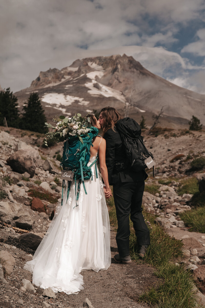 Adventure elopement couple in wedding attire stands on a mountain trail with snowy Mount Hood in the background, capturing a scenic Oregon alpine moment.