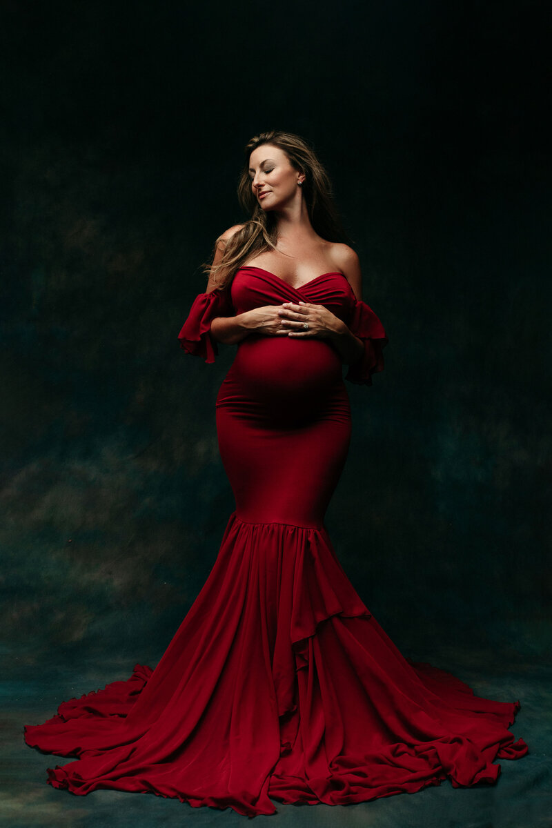 High end pregnancy portrait of 35 week pregnant lady wearing a beautiful off the shoulder maroon  fitted gown  holding hands on top of her tummy and closing eyes head to the side
