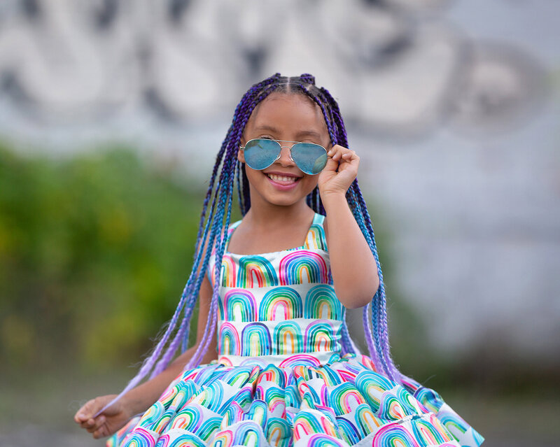 A little girl with long blue and purple braids is holding her sunglasses and smiling. She's wearing a rainbow tutu dress and standing in front of a wall with graffiti.