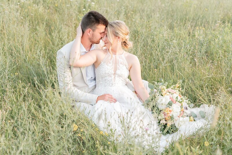 A bride sits on her groom's lap in a summer field, foreheads touching.