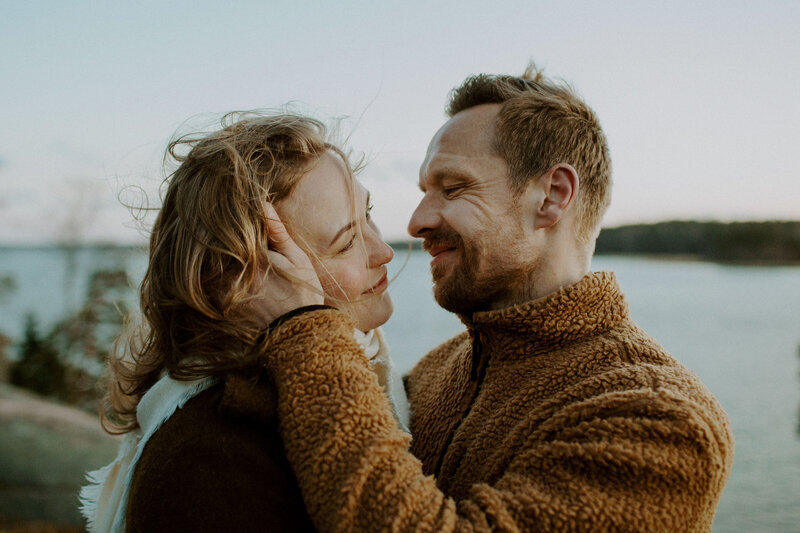 Engaged couple standing at the cliff looking each other smiling and he is touching her face with his hand in Espoo in Finland