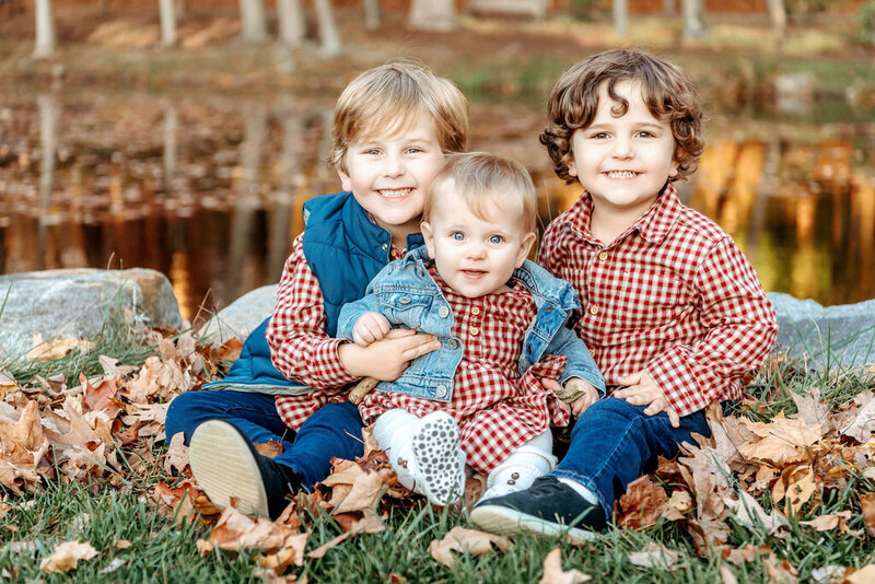 Three young siblings sitting together in autumn leaves, wearing coordinating gingham shirts and denim, with the older children lovingly embracing their baby sibling in a natural outdoor setting