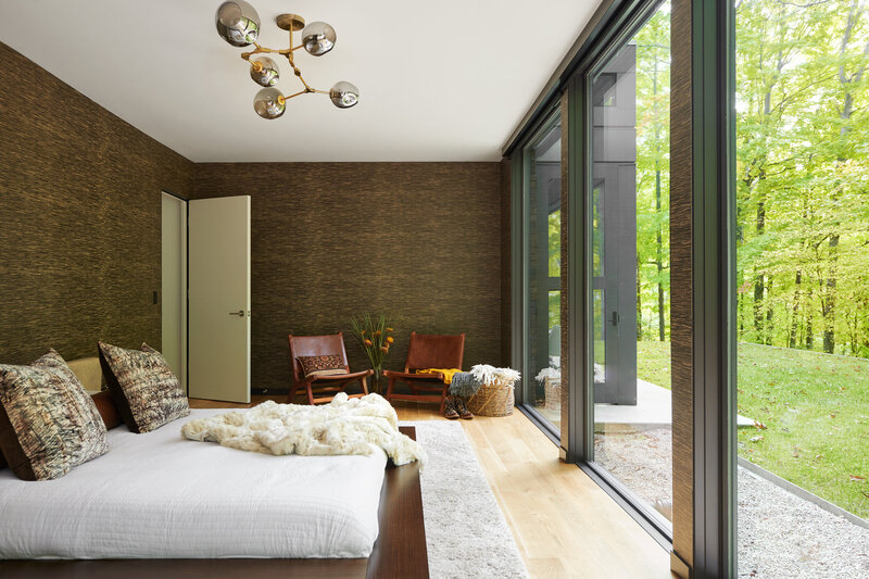 Master Bedroom with dark wallpaper, large windows,  a low modern bed and leather chairs. The lighting is a mix of soft gold and silver antiqued glass. The dark walls are a black and neutral grass cloth wallpaper. Wood floors are white oak.