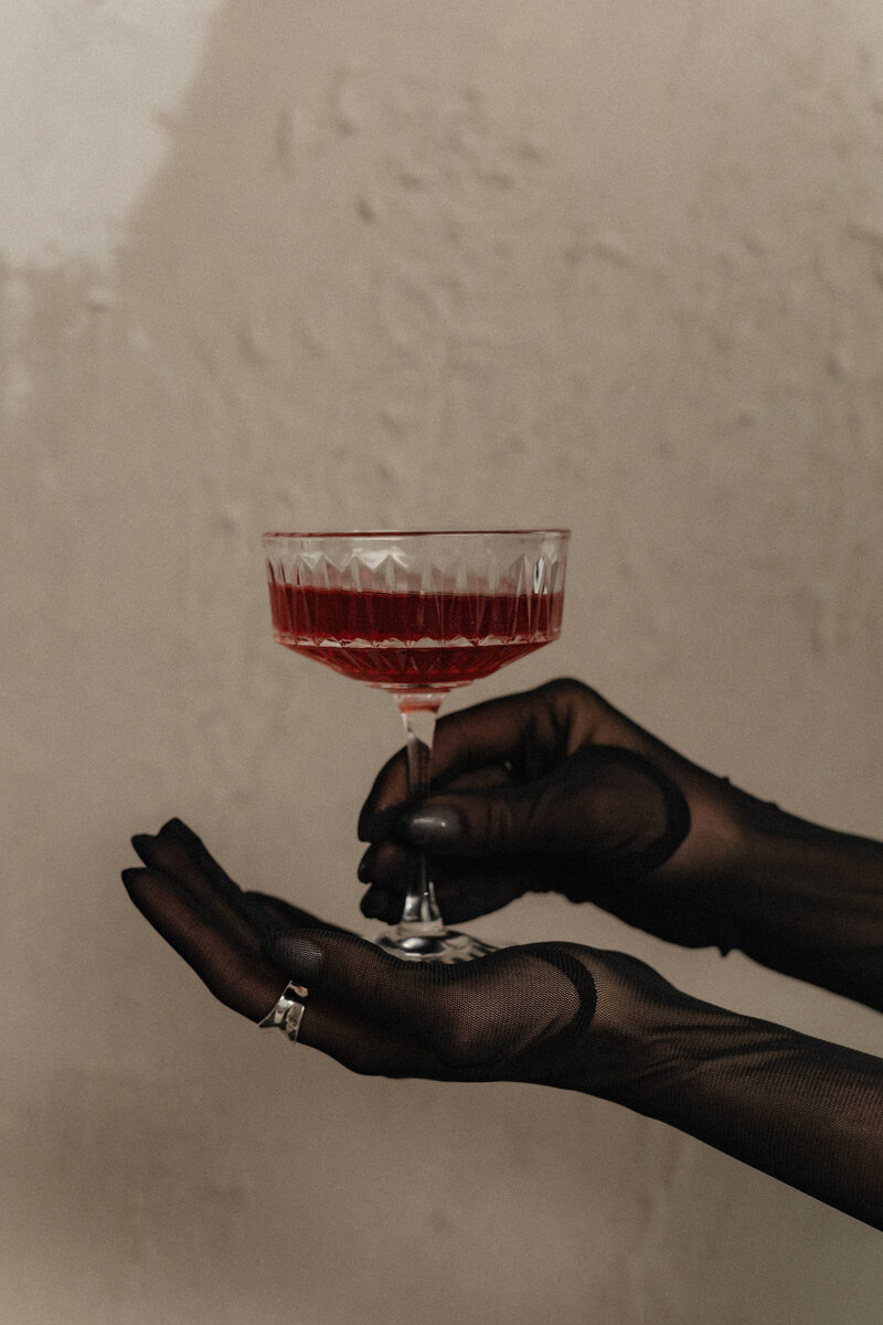 A womans hand covered in black gloves balancing a glass with a red drink while using her other hand covered in black gloves to hold the stem of the glass.