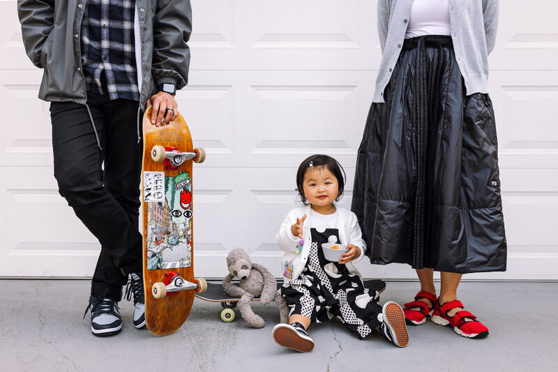 toddler girl with snack sitting on skateboard between two parents in front of garage door