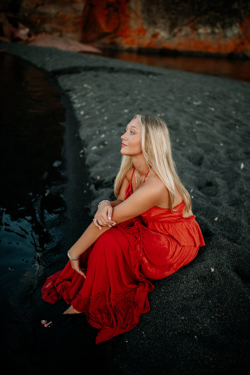 High school senior wearing a vibrant red maxi dress sits along Black Beach in Silver Bay during a summer senior session.